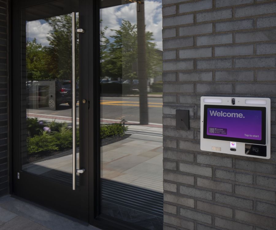Cadillac Manor Touchscreen kiosk with "Welcome" message is mounted beside a glass door on a modern brick building.