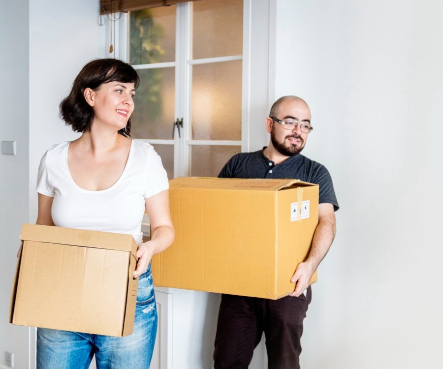Cadillac Manor Two people smiling and carrying cardboard boxes inside a bright room.