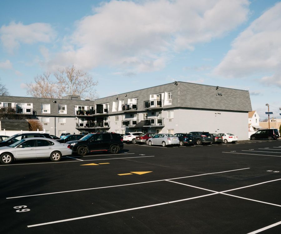 Cadillac Manor A parking lot with several parked cars in front of a gray apartment building under a partly cloudy sky.