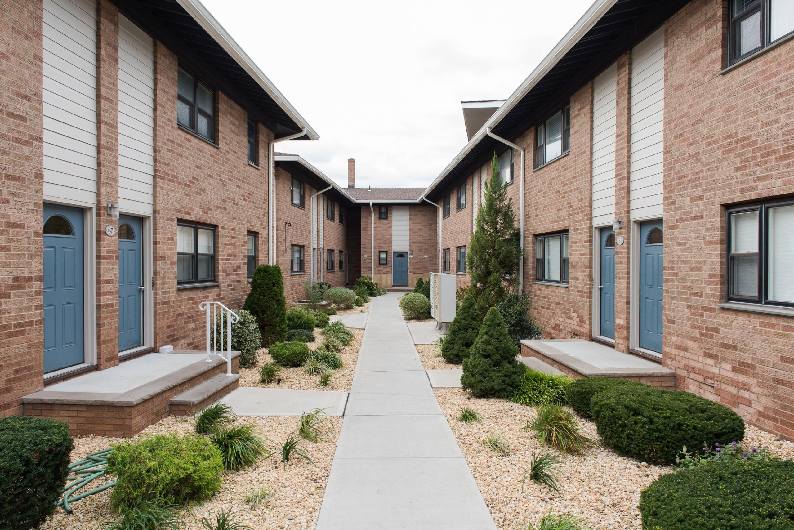 Cambridge Manor Two gray armchairs with pillows sit by a small table in a modern, gray-toned hallway near an elevator.