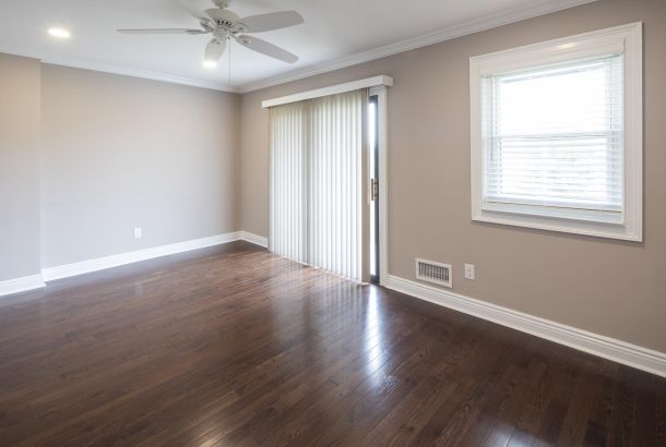 Cambridge Manor Empty room with hardwood floors, beige walls, ceiling fan, window, and sliding glass door with vertical blinds.