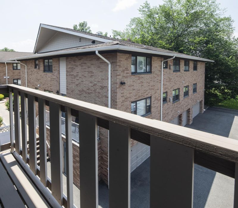 View from a balcony overlooking a brick apartment building and a parking area with trees in the background.