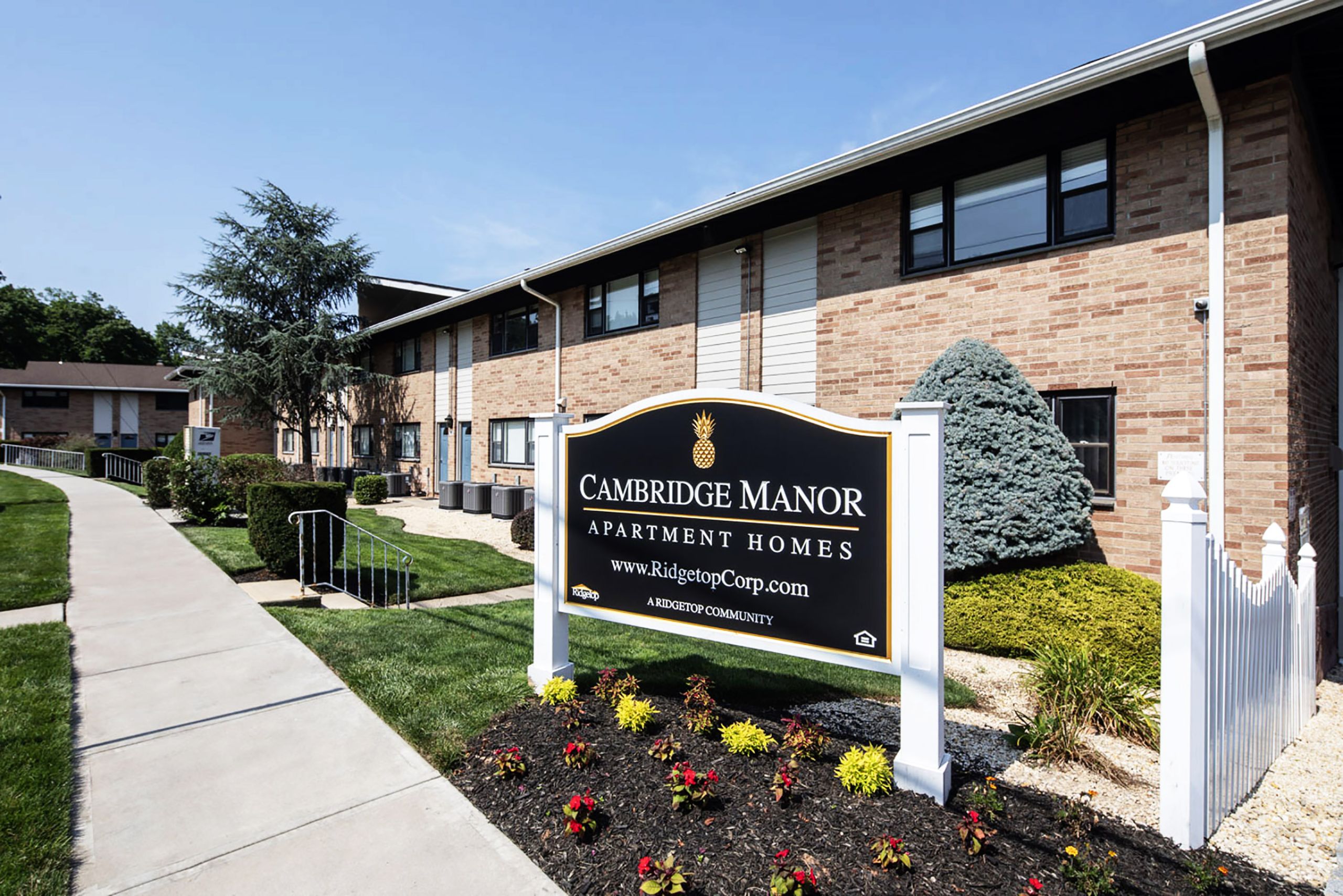 Cambridge Manor A modern apartment building with landscaped gardens and a wooden entrance canopy under a partly cloudy sky.