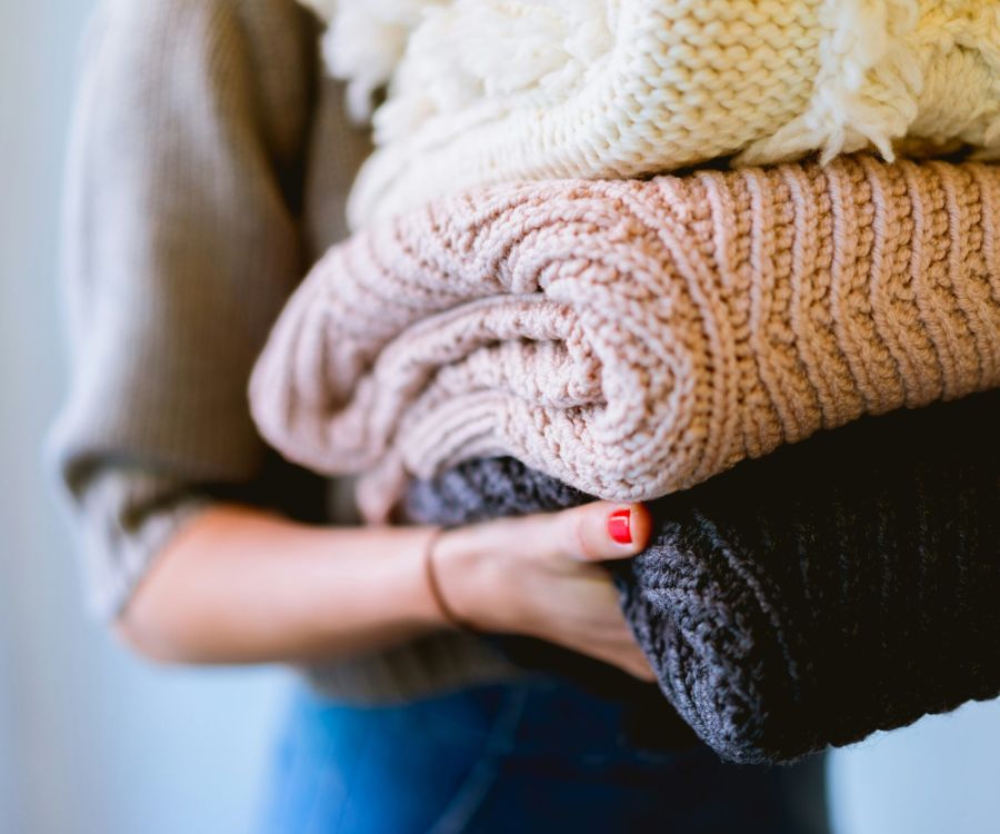 Person holding a stack of folded knit sweaters in neutral colors.