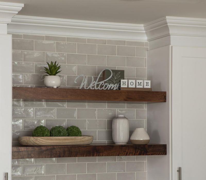 Two wooden shelves with decor, including plants, vases, and a "Welcome Home" sign, against a tiled wall.