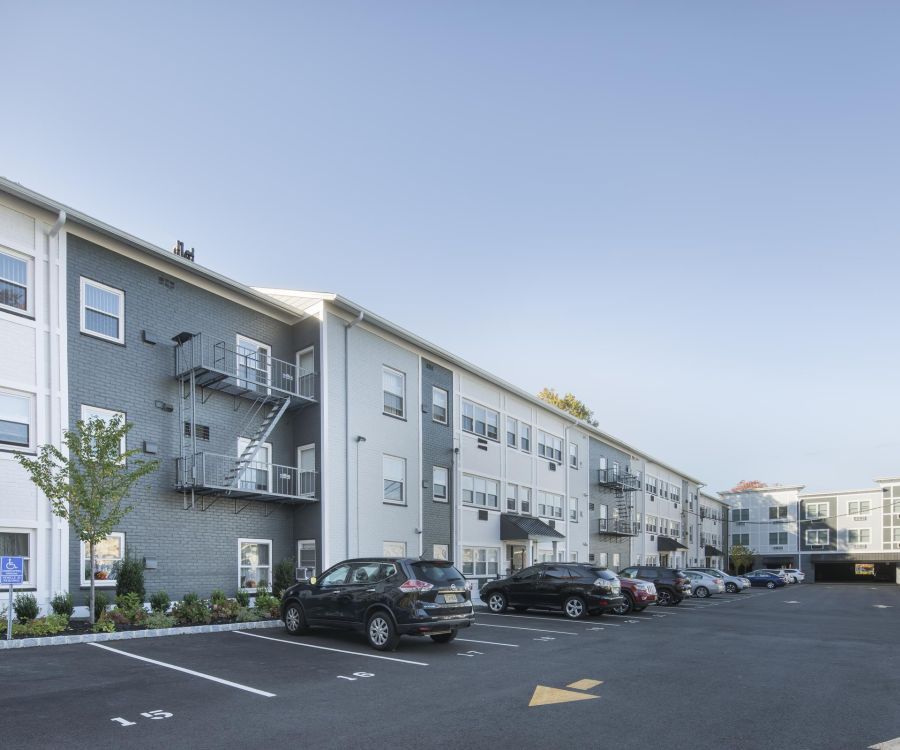 Hickory Manor Wide view of a modern apartment building with parked cars in a lot on a clear, sunny day.