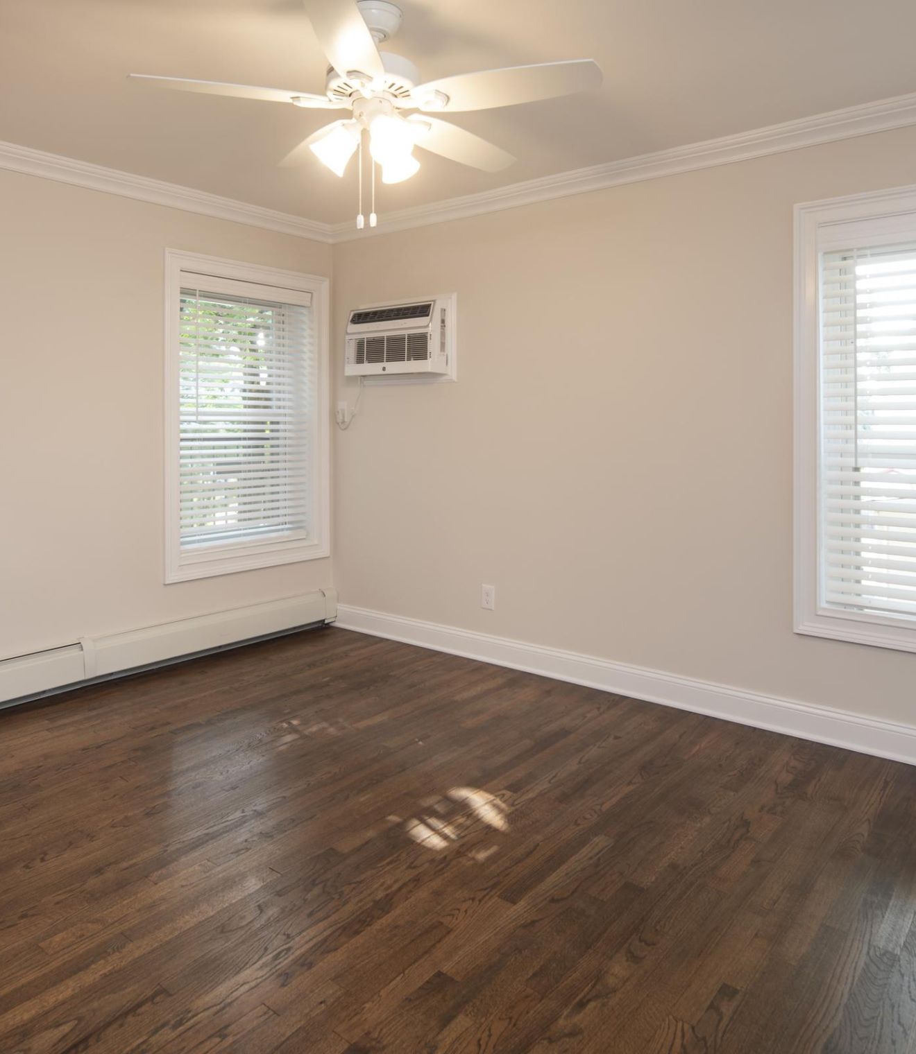 Hickory Manor Empty room with hardwood floor, two windows with blinds, ceiling fan, and wall-mounted air conditioner.