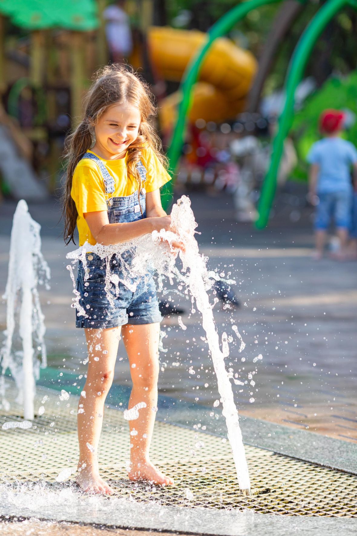 Hickory Manor Smiling girl in denim overalls plays with water fountain at a sunny outdoor playground.