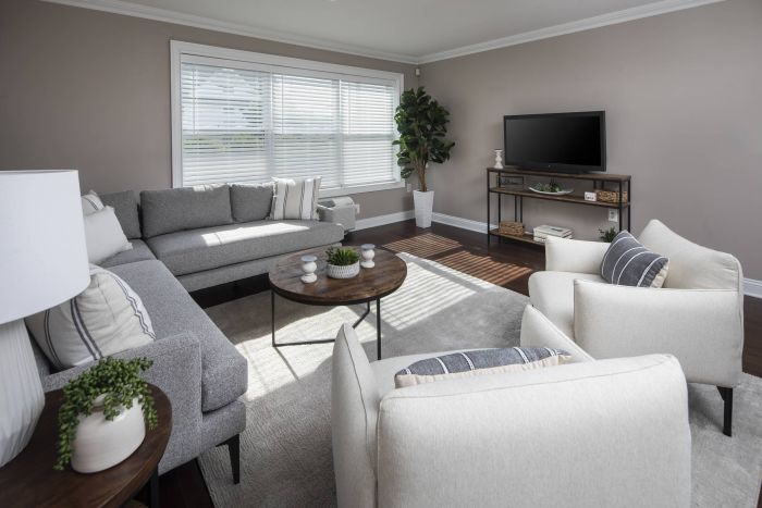 Modern living room with gray sofa, two beige chairs, a round coffee table, TV, and large window with blinds.
