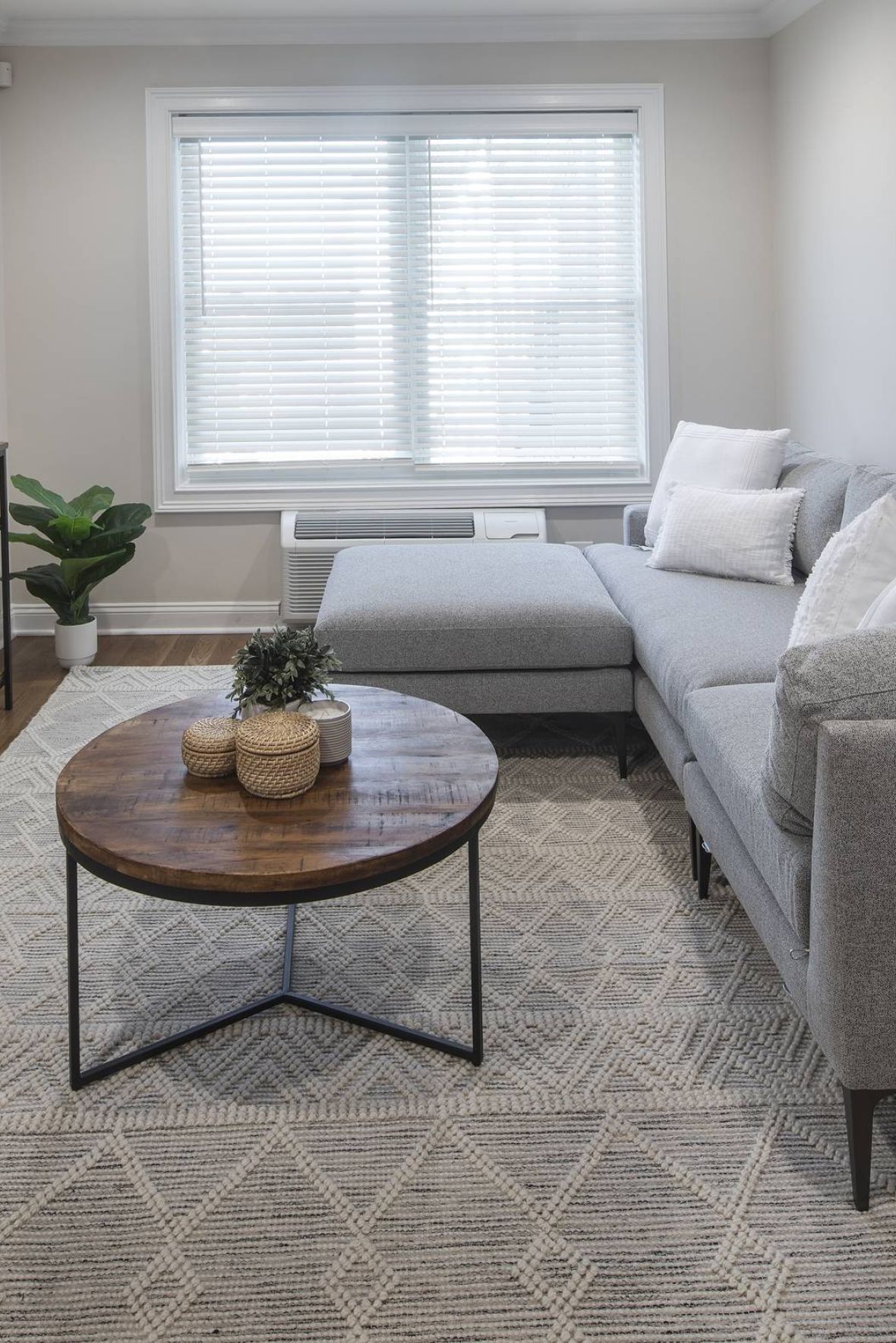 Modern living room with a grey sectional sofa, round wooden coffee table, TV, and large window with blinds.