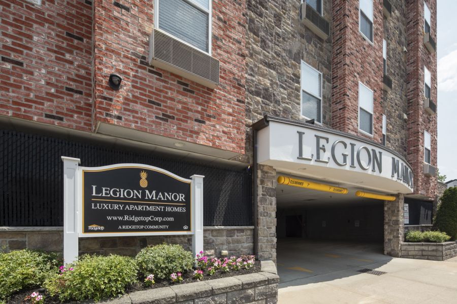 Legion Manor A modern apartment building with landscaped gardens and a wooden entrance canopy under a partly cloudy sky.
