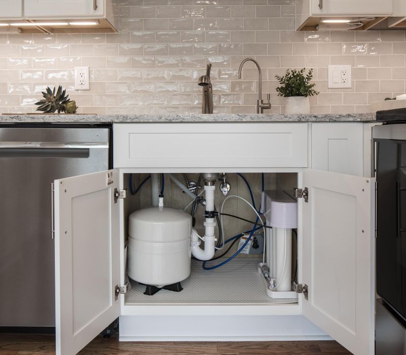 Under-sink reverse osmosis water filtration system in a modern white kitchen with natural wood floors and shelving.