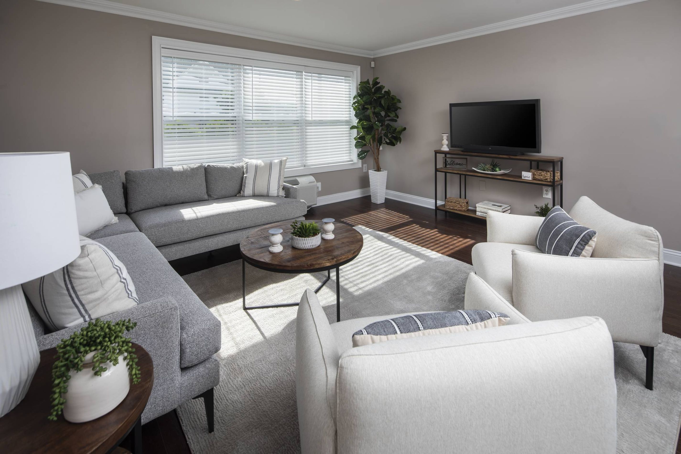 Modern living room with gray sofa, two beige chairs, a round coffee table, TV, and large window with blinds.