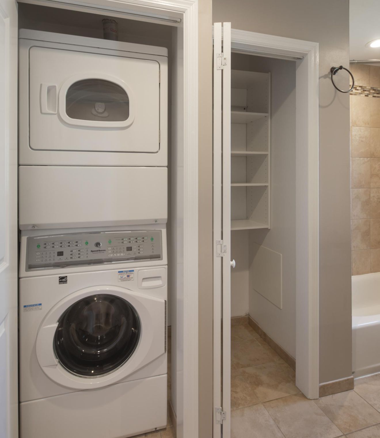 Stacked washer and dryer next to an open closet in a tiled laundry area with beige walls.
