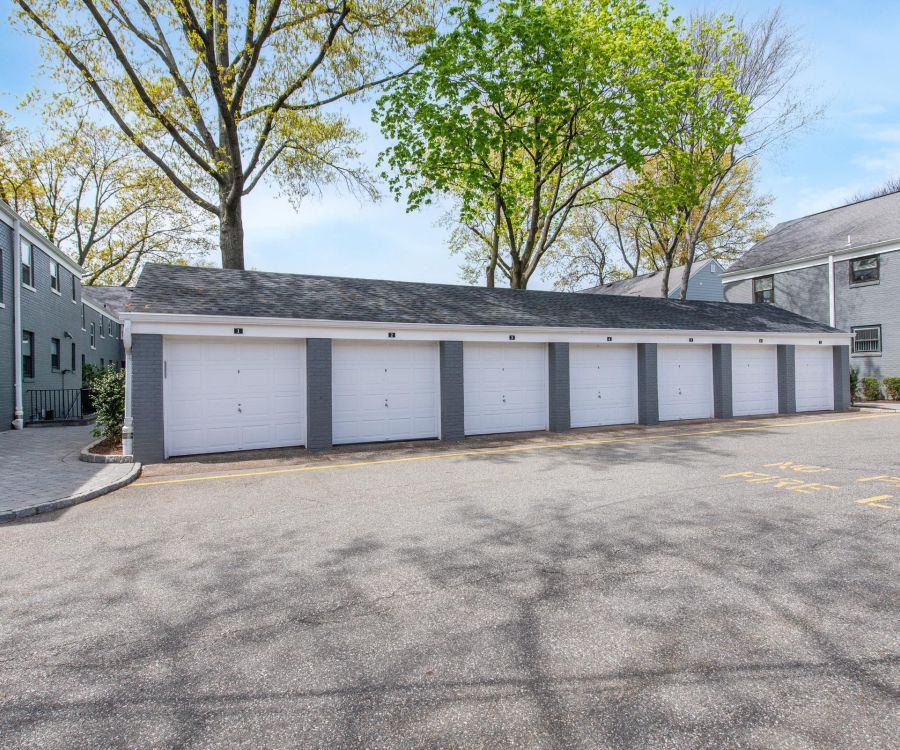 A row of six closed white garage doors with gray trim beside two apartment buildings and trees.