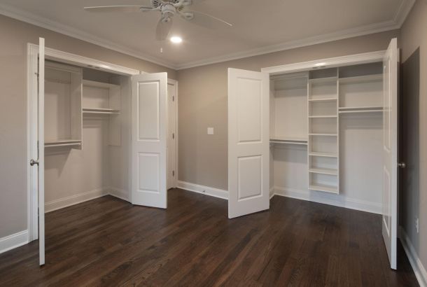 Empty bedroom with dark wood floors, open double closets, beige walls, and ceiling fan with lights.