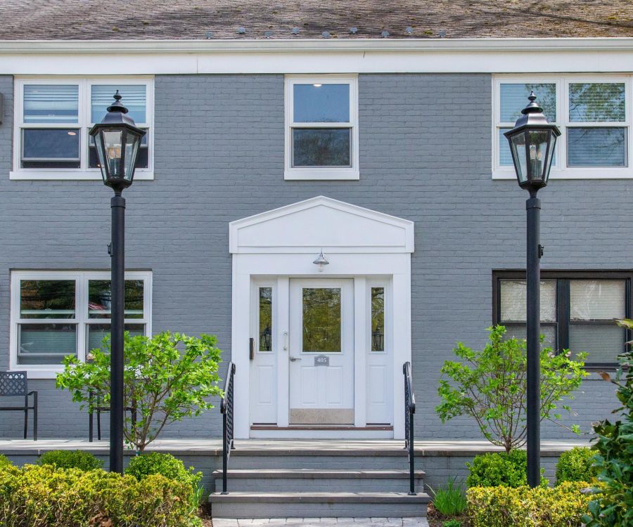 Gray-brick building with white double doors, black lamps, and green bushes at the entrance.