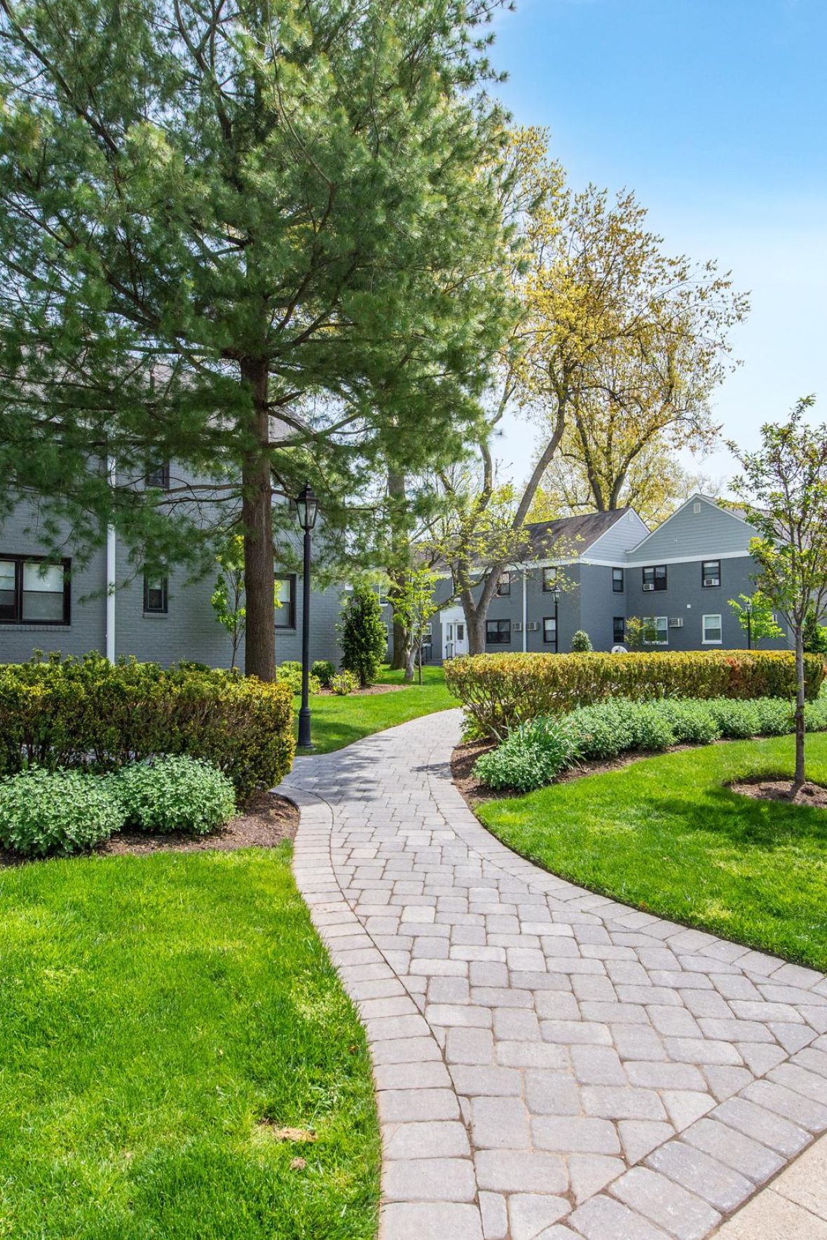 Leonia Manor Empty outdoor tennis court with nets, surrounded by trees and buildings, under a partly cloudy sky.