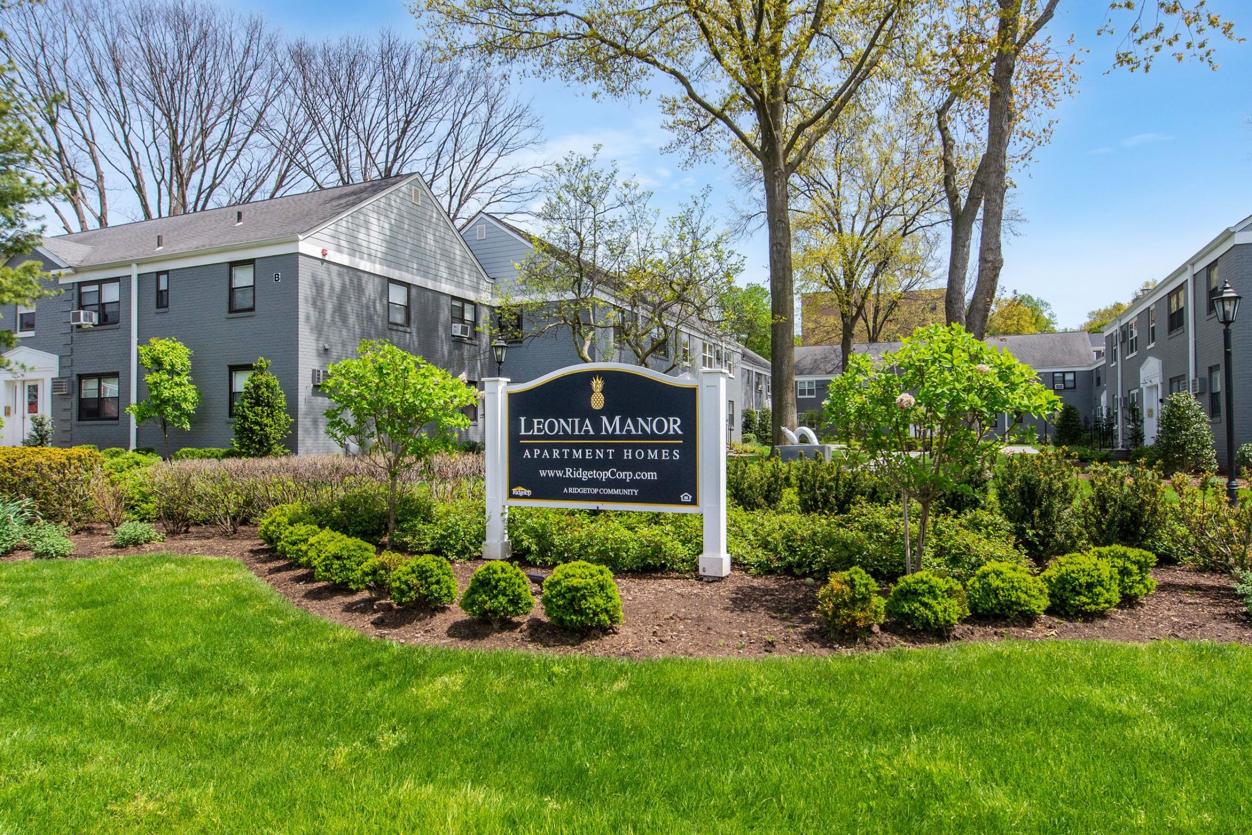 Leonia Manor A modern apartment building with landscaped gardens and a wooden entrance canopy under a partly cloudy sky.