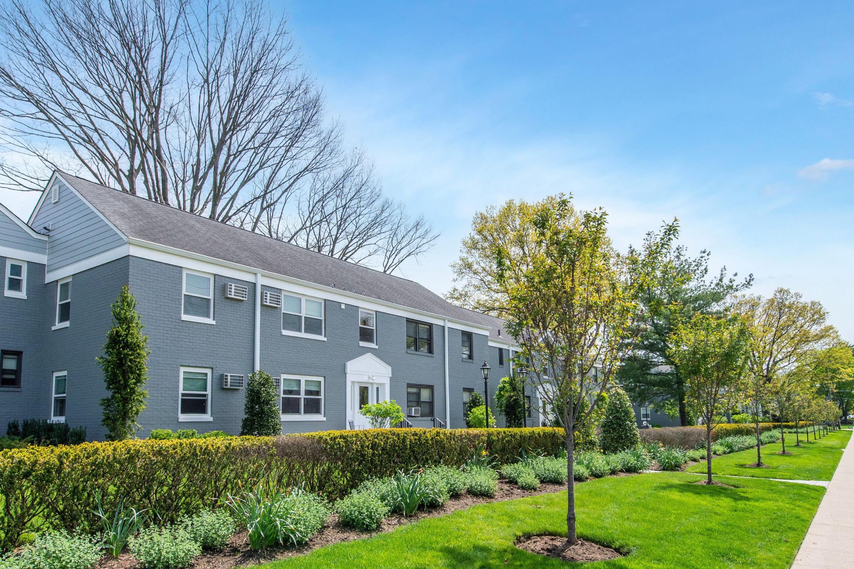 Gray apartment building with white trim, surrounded by green lawns, bushes, and trees on a sunny day.
