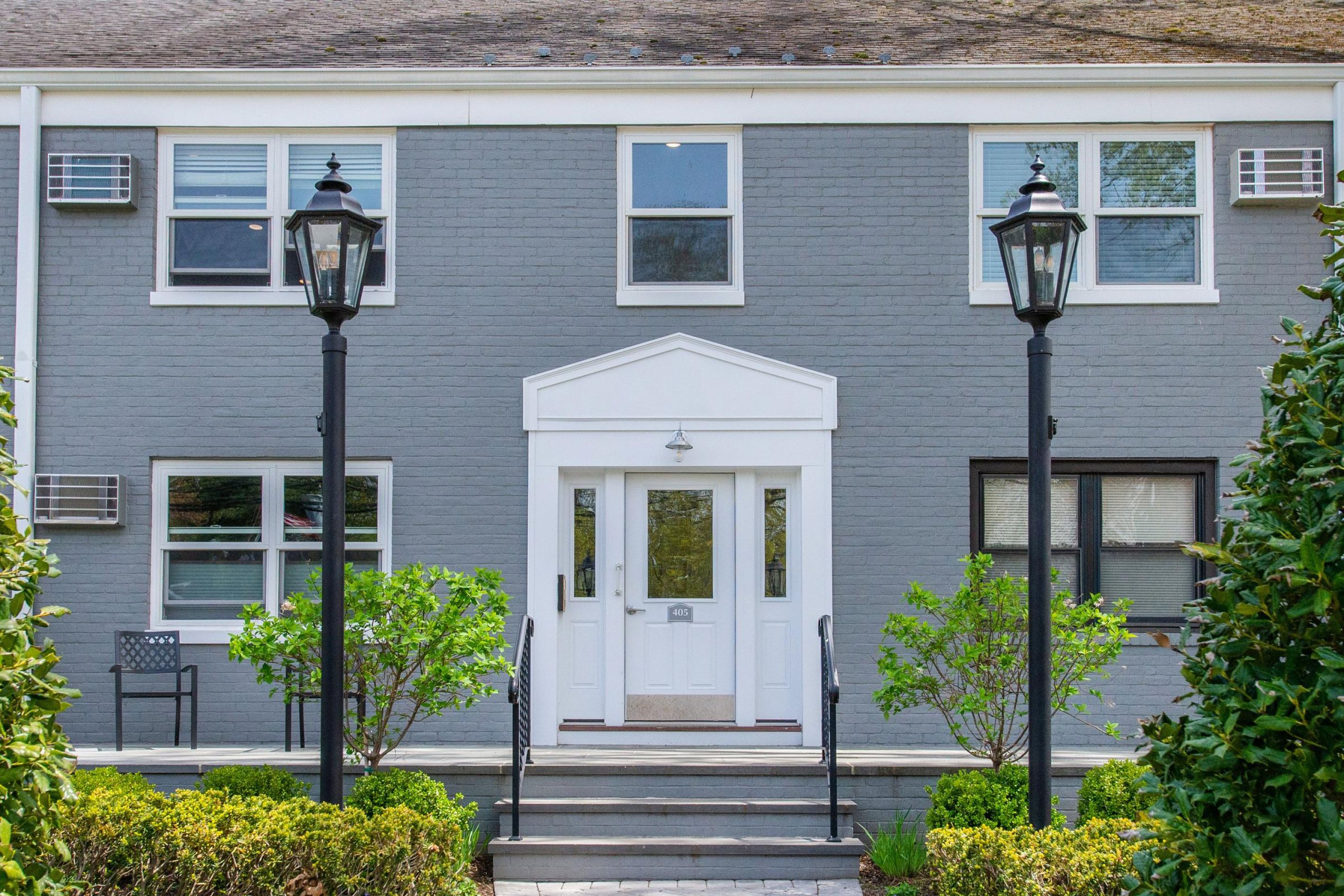 Gray-brick building with white double doors, black lamps, and green bushes at the entrance.