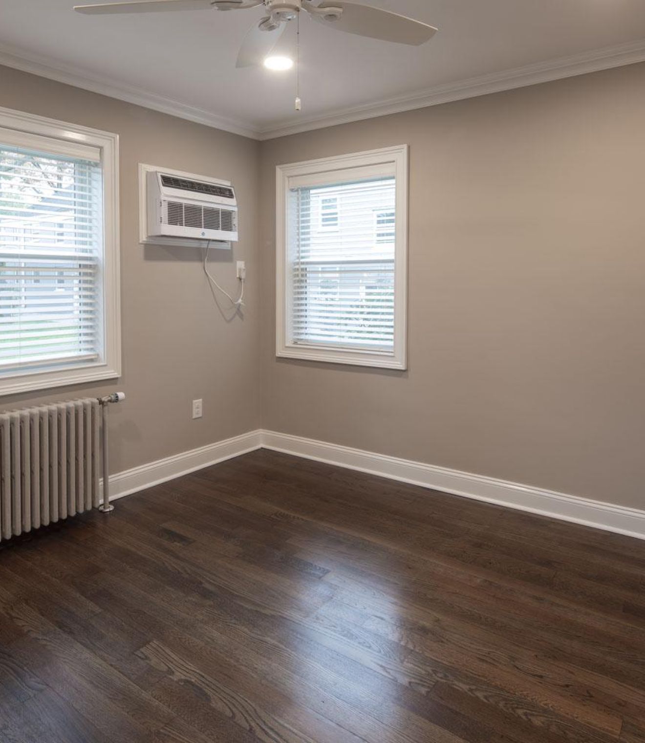 Empty room with hardwood floors, two windows, ceiling fan, radiator, and window air conditioner.