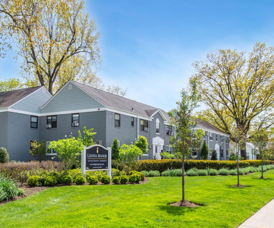 Modern apartment buildings with gray siding, green lawns, trees, and a sign reading "Leonia Manor" in front.