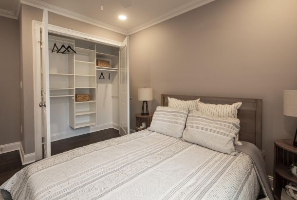 A neatly made bed faces an open walk-in closet with shelves, baskets, hangers, and bedside lamps.