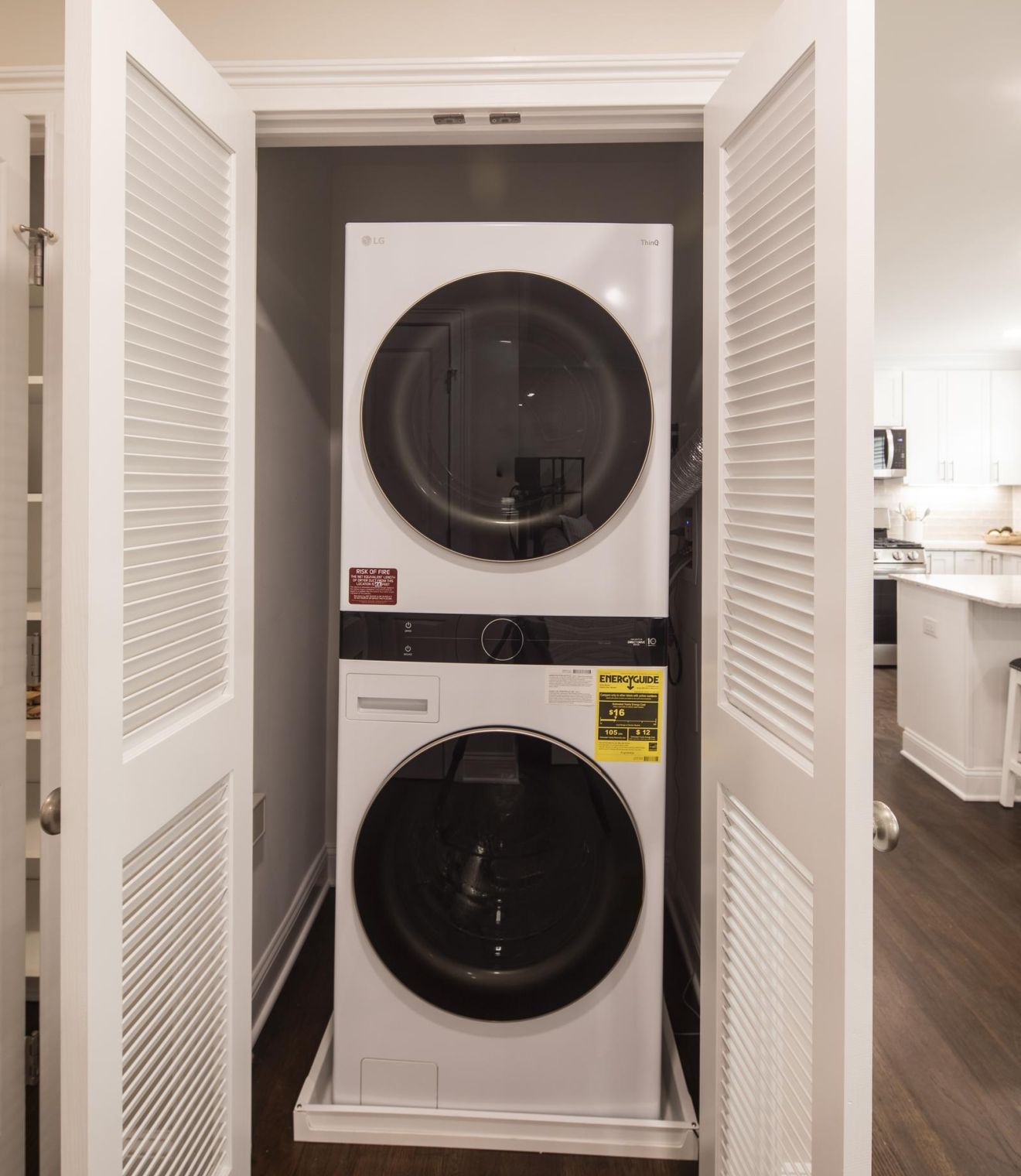 Stacked washer and dryer in a small laundry room with white walls, wooden floor, and open shelving.