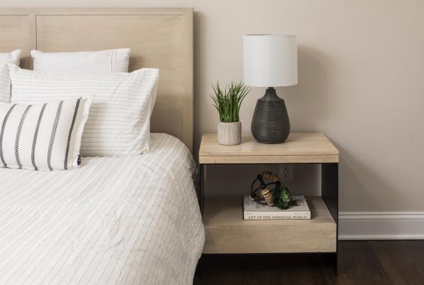 A bed with white linens next to a wooden nightstand with a lamp, plant, and books.