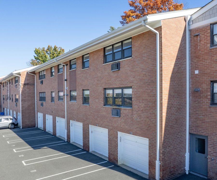 Brick apartment building with garage doors, parked cars, and empty parking spaces on a sunny day.