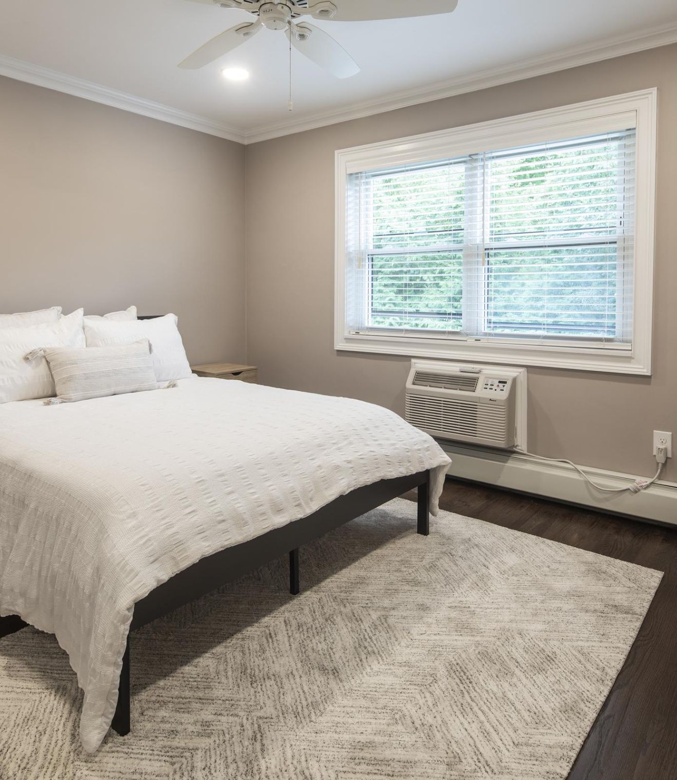 Bedroom with a neatly made bed, armchair, window, ceiling fan, and neutral-colored walls and decor.
