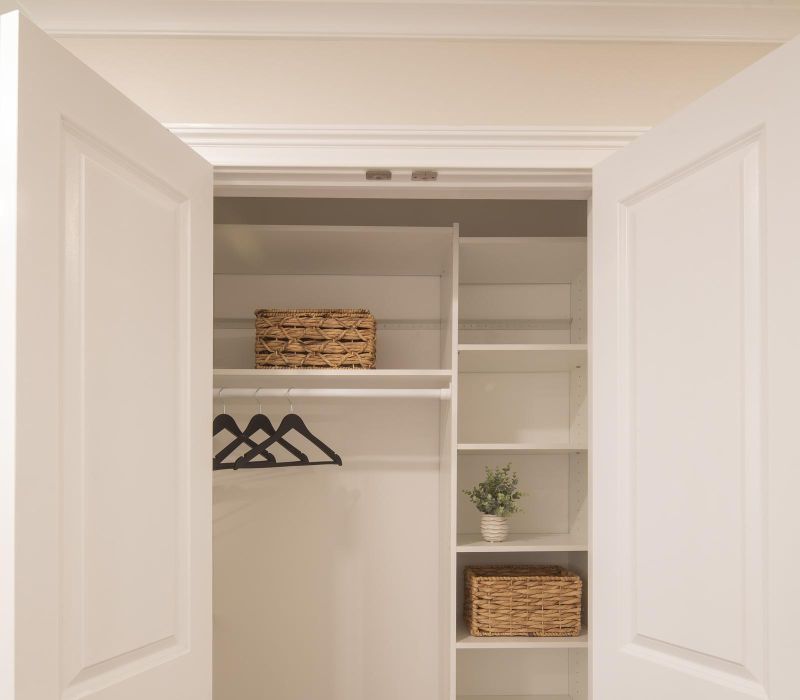 Open white closet doors revealing shelves, baskets, hangers, and a small potted plant inside a tidy closet.