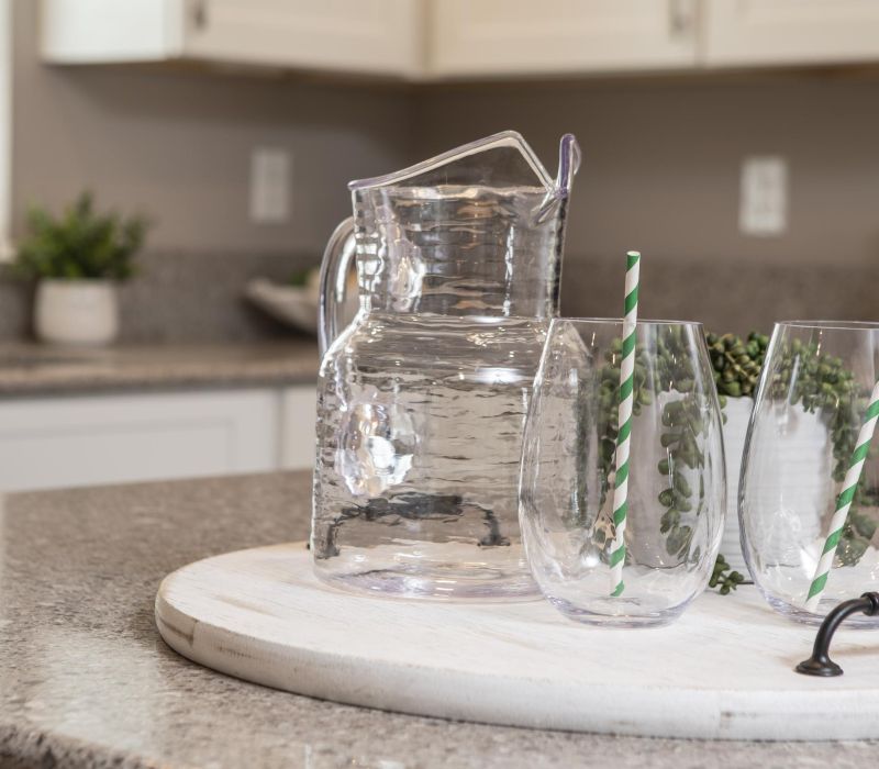 Clear glass pitcher and two glasses with green striped straws on a white tray on a kitchen counter.