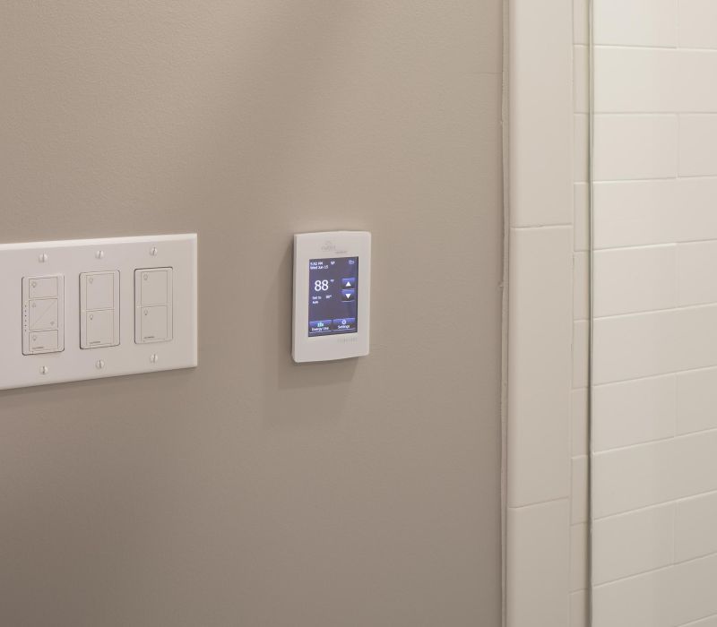 A smart thermostat on a beige wall next to light switches, near white tiled bathroom walls.
