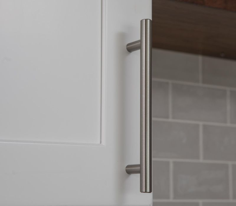 Close-up of a silver metal handle on a white cabinet door with gray tiled backsplash in the background.