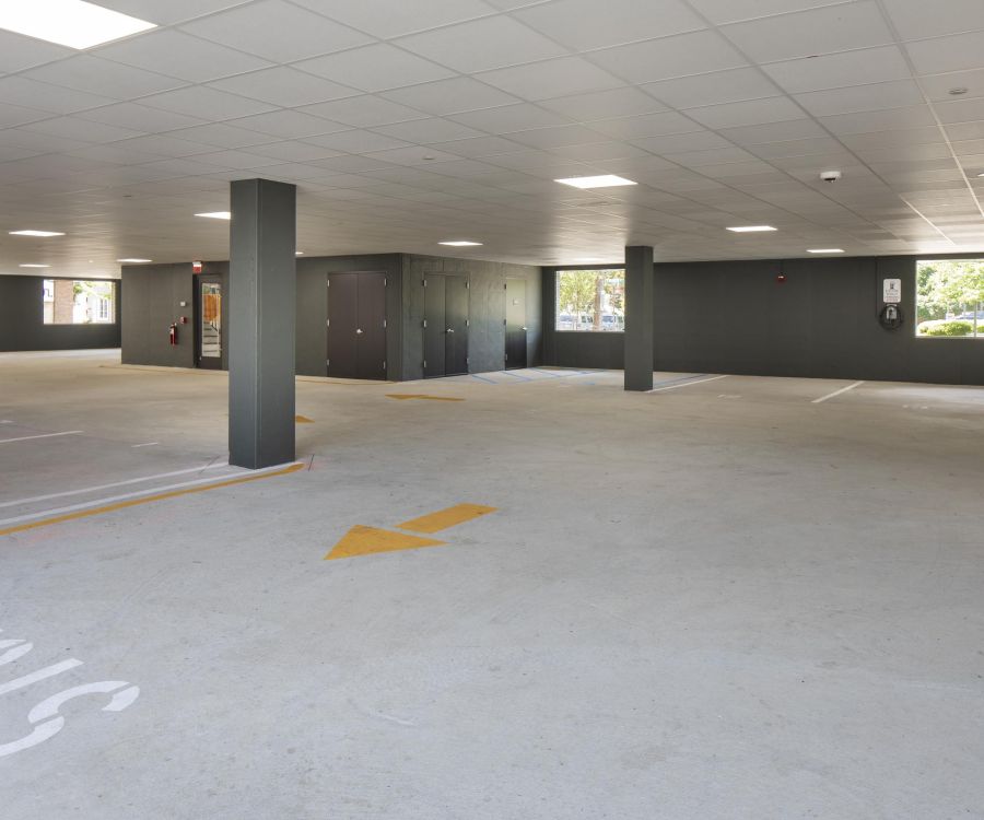 Empty indoor parking garage with gray pillars, yellow arrows on the floor, and sunlight through windows.