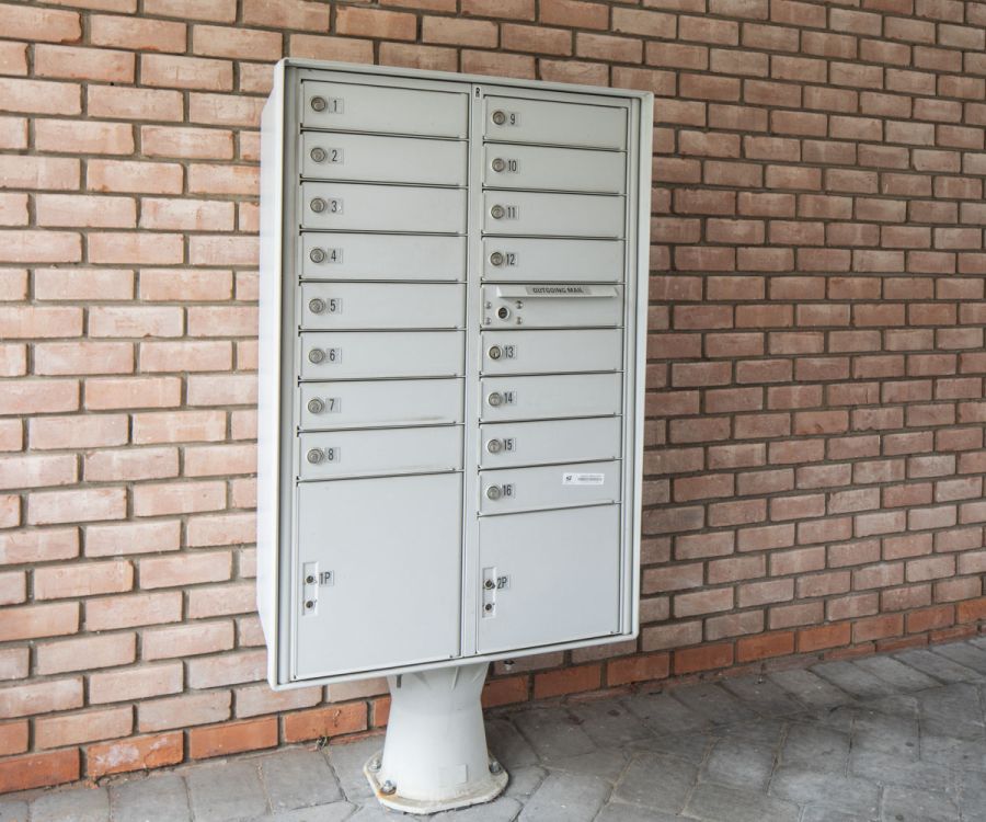 A gray metal mailbox unit with multiple compartments stands against a red brick wall.