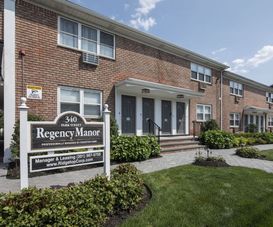 Brick apartment building with a Regency Manor leasing sign on a well-kept lawn under a sunny sky.