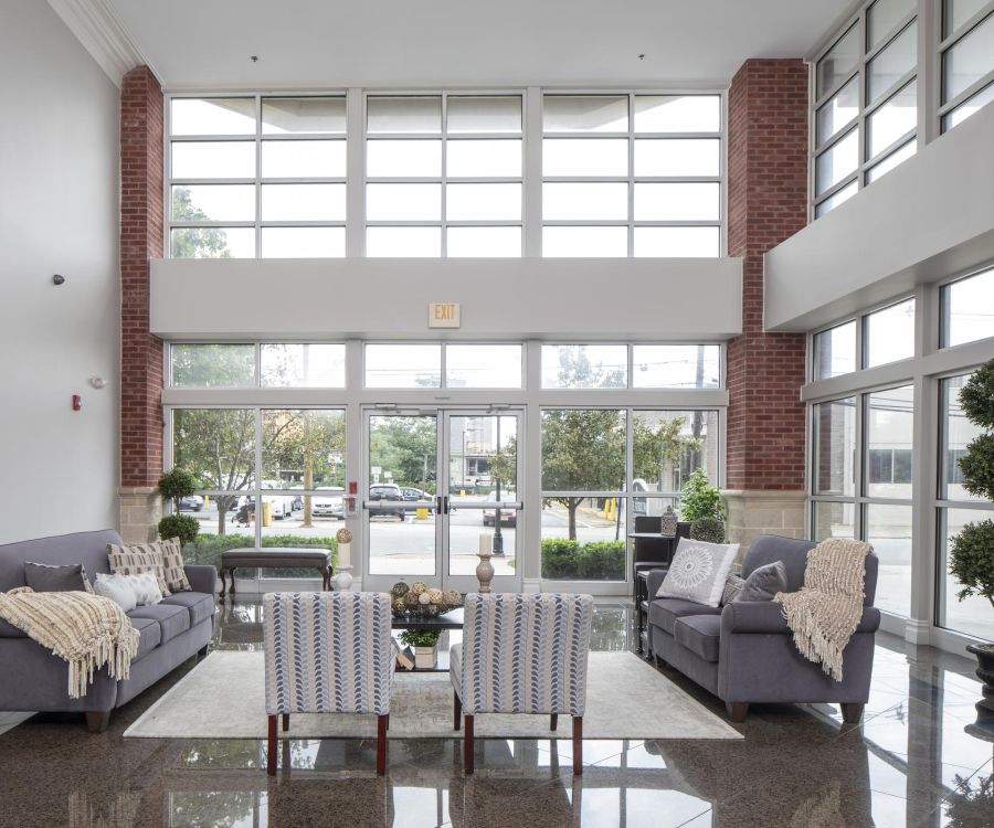 The Atrium Modern lobby with large windows, two sofas, armchairs, and coffee tables, featuring neutral decor and greenery.