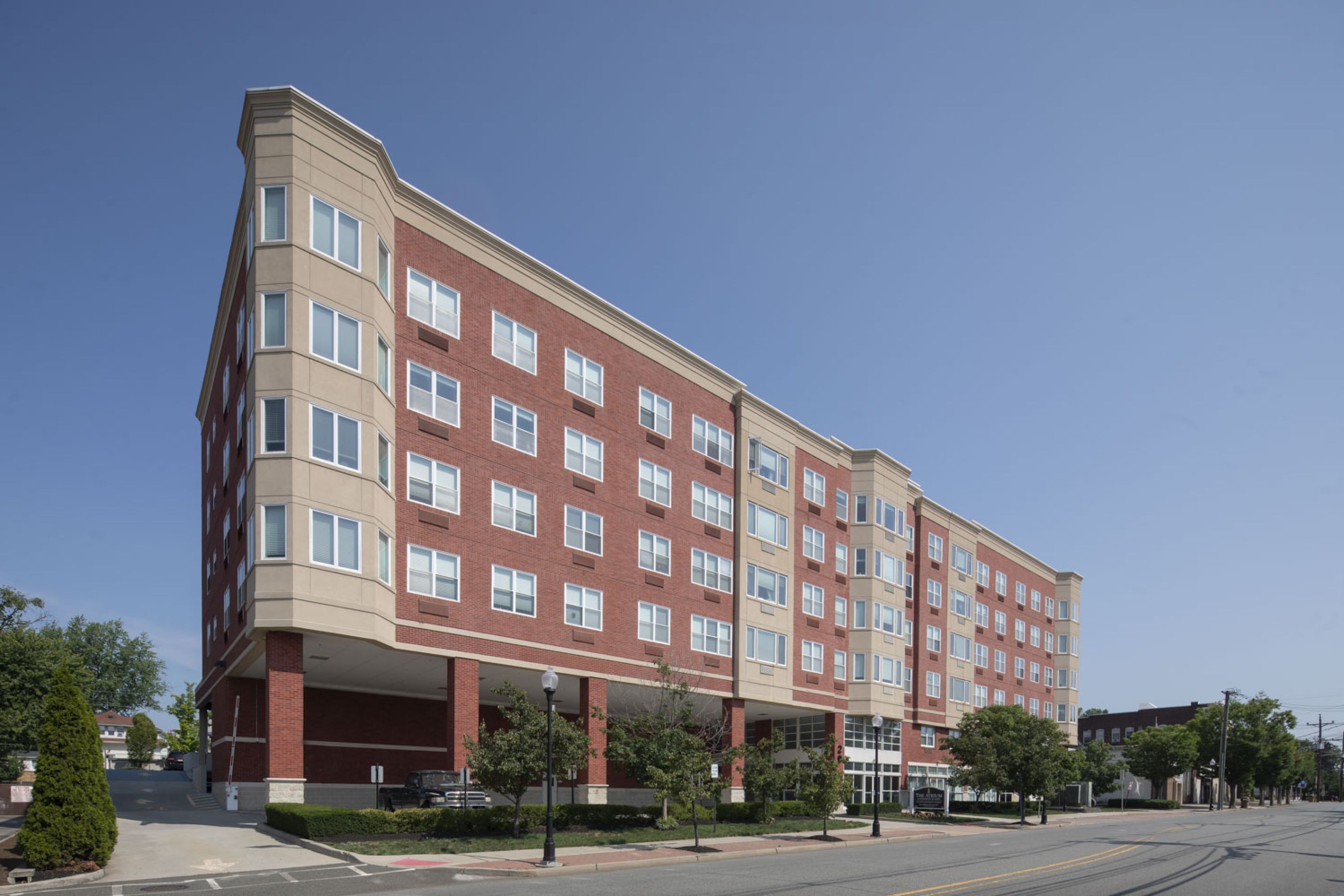 The Atrium A modern, mid-rise red brick apartment building on a city street under a clear blue sky.