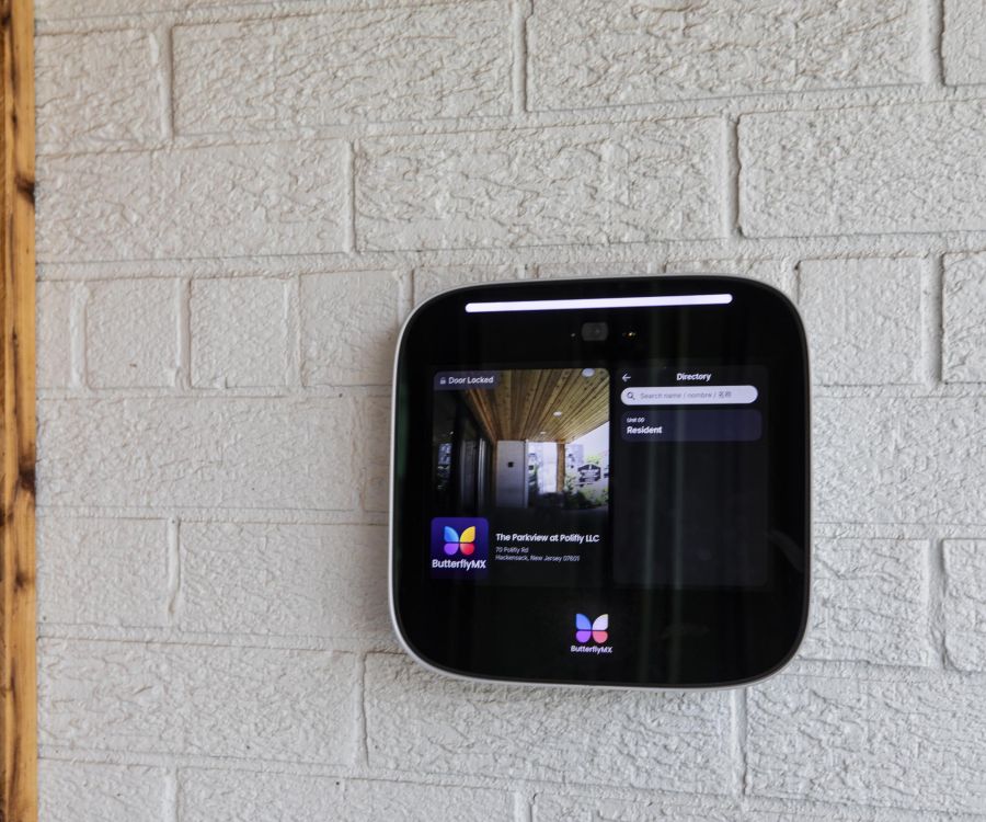 A digital ButterflyMX intercom system is mounted on a white brick wall next to a wooden corner.