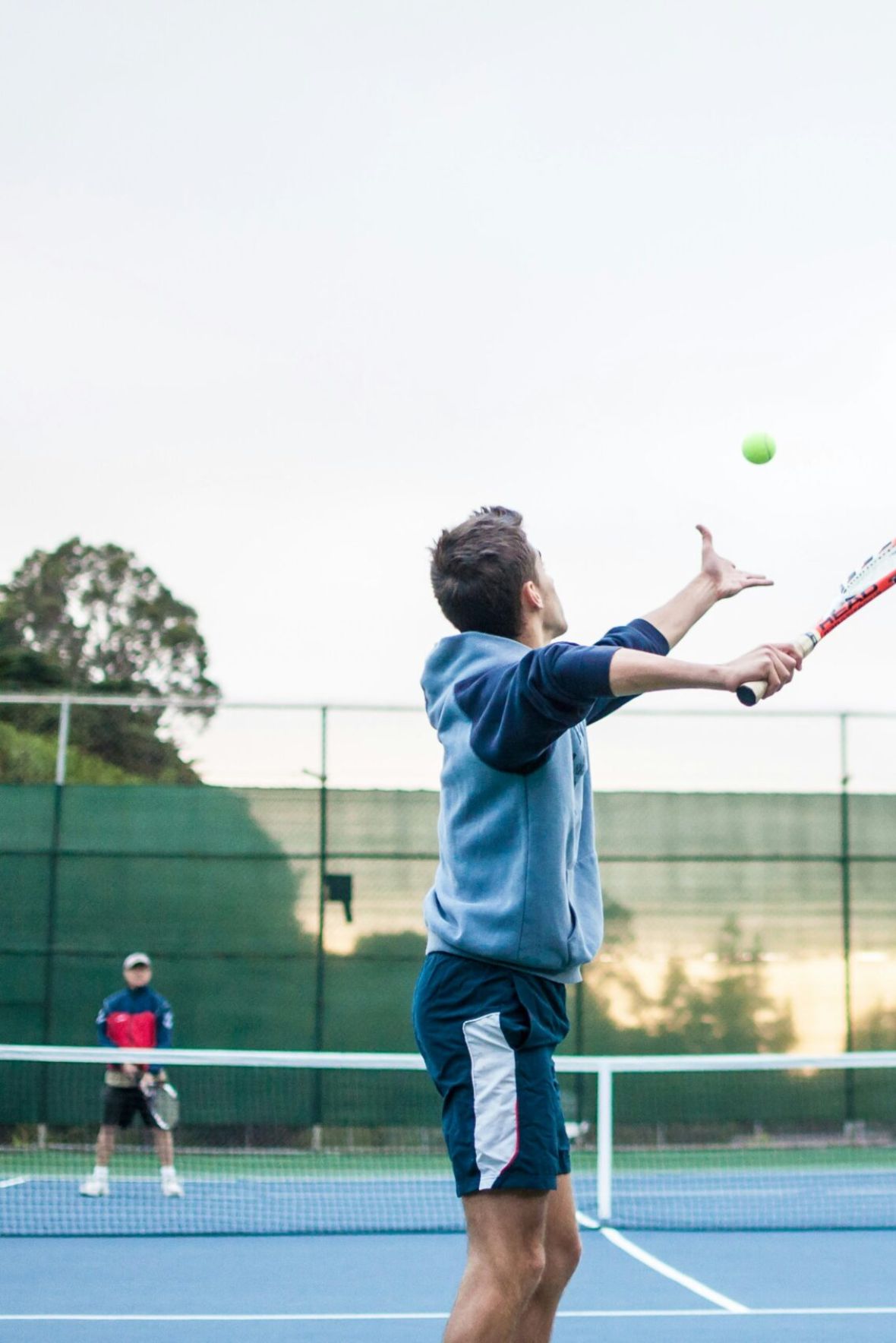 The Cornerstone Empty outdoor tennis court with nets, surrounded by trees and buildings, under a partly cloudy sky.