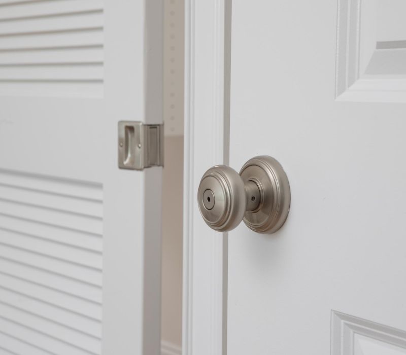 Close-up of a white door with a round silver doorknob next to an open louvered closet door.