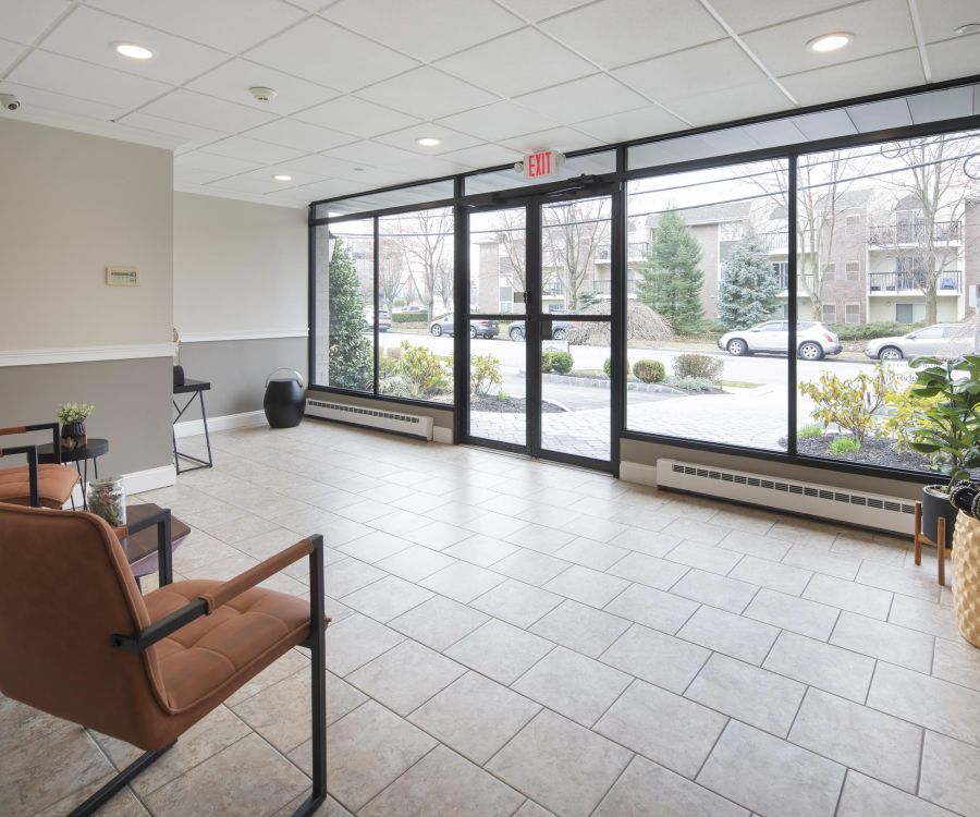 Modern lobby with large windows, brown chairs, tiled floor, and plants by the entrance.