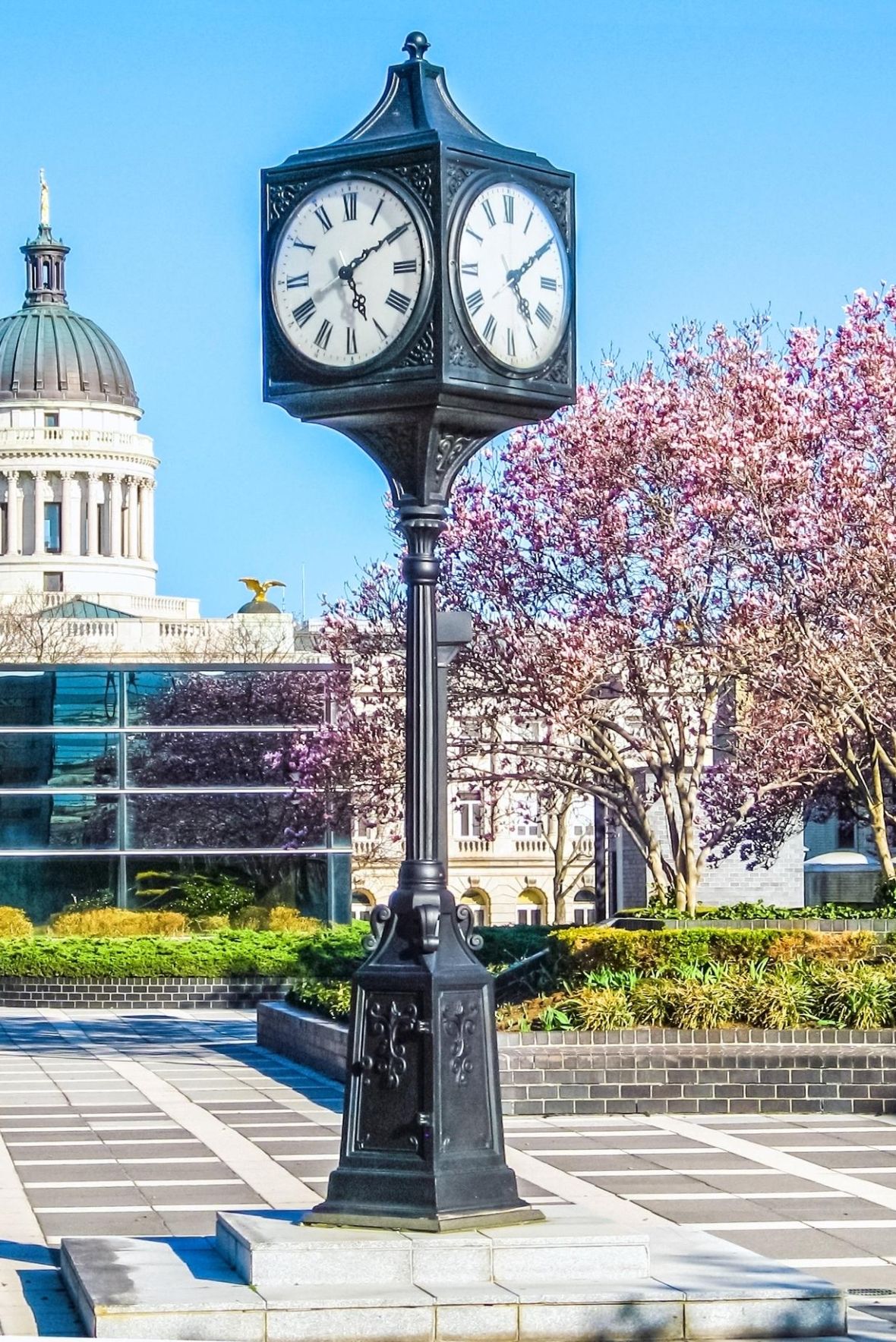 The Cornerstone A vintage clock stands in a plaza with blooming trees and a domed building in the background.