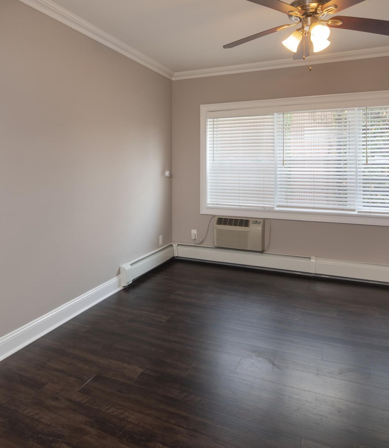 Empty room with dark wood floor, beige walls, large window with blinds, and ceiling fan with lights.