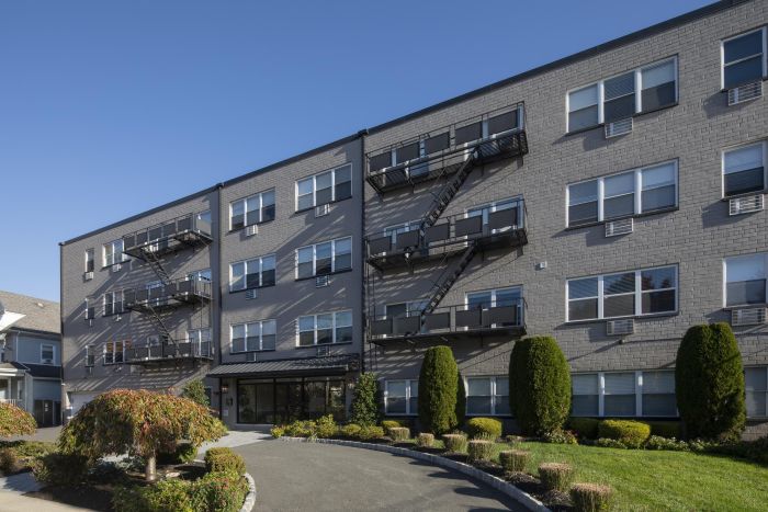 A modern four-story brick apartment building with fire escapes and neatly trimmed bushes in front.