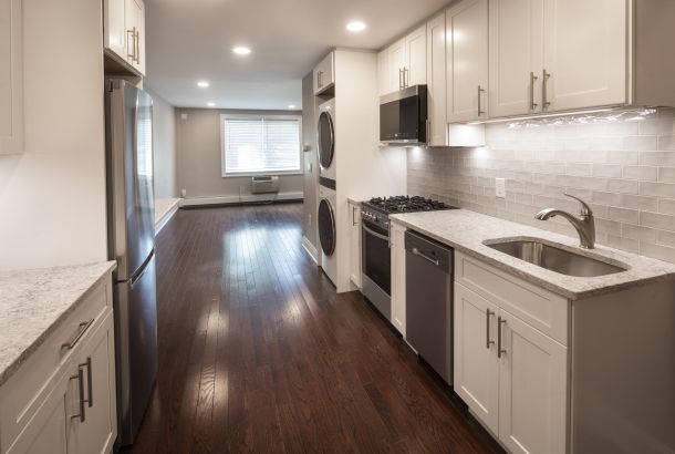 Modern kitchen with white cabinets, stainless steel appliances, and dark wood flooring leading to a bright living area.