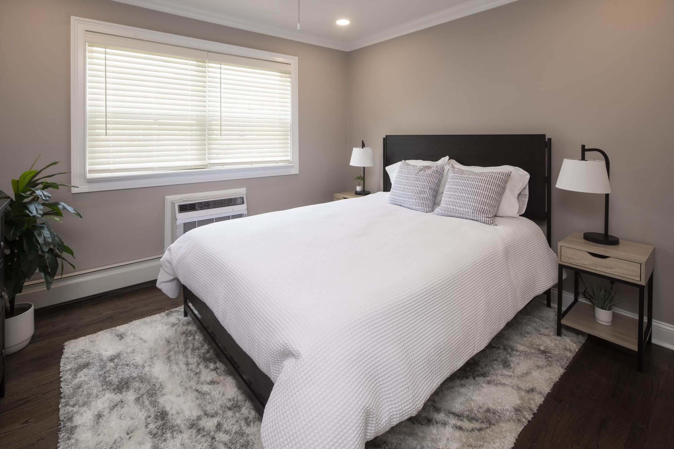 A neatly made bed in a modern bedroom with gray walls, white bedding, and natural light from a window.